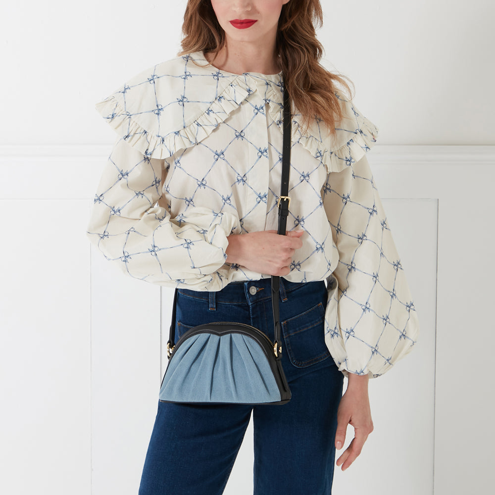 A woman in a cream blouse with blue bows and dark-wash jeans is modelling a half-moon shaped denim crossbody bag with black leather trim.