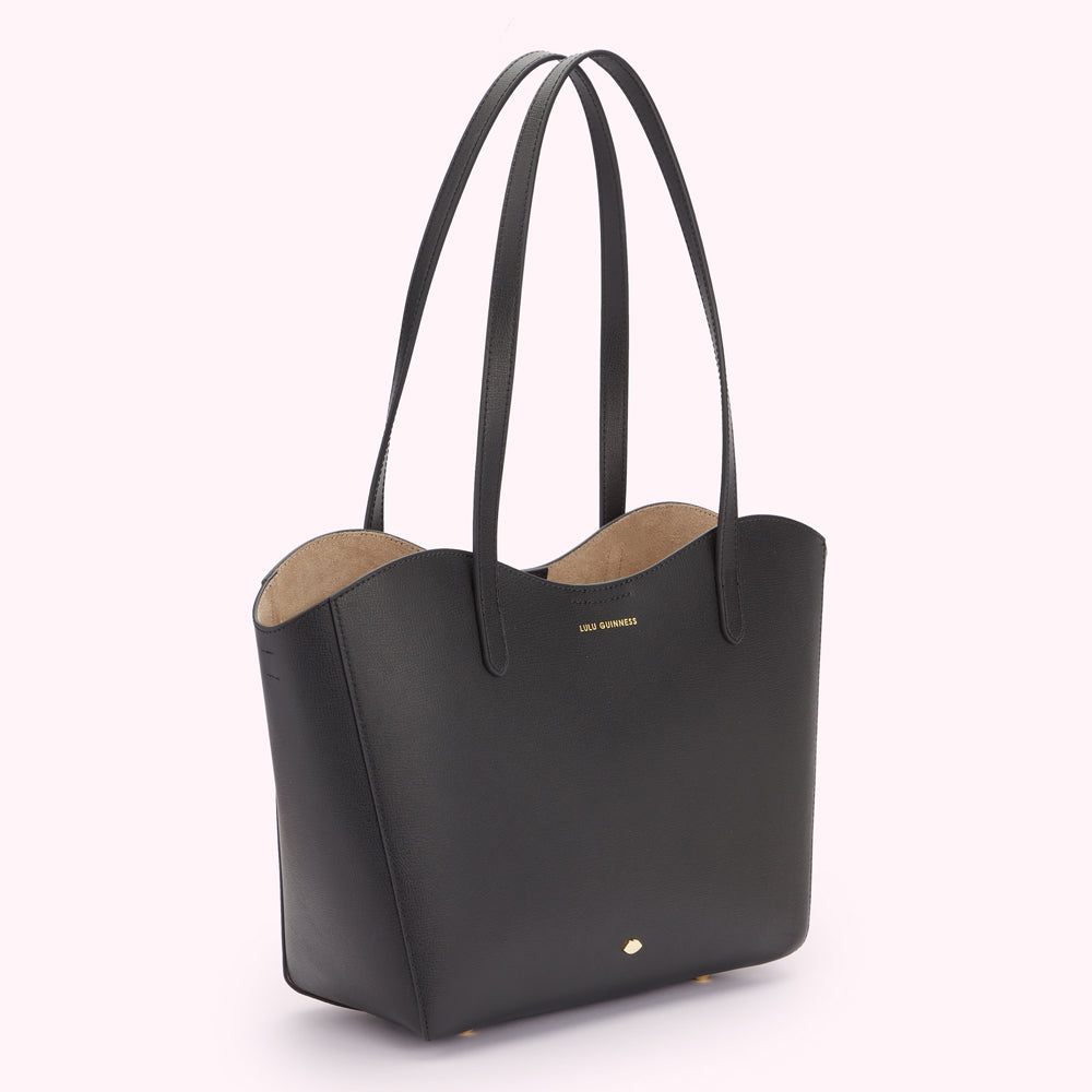 Angle image of a black leather tote bag with a curved top edge. Showing the depth of the bag and the gold lip pin and foil print Lulu Guinness logo detail.