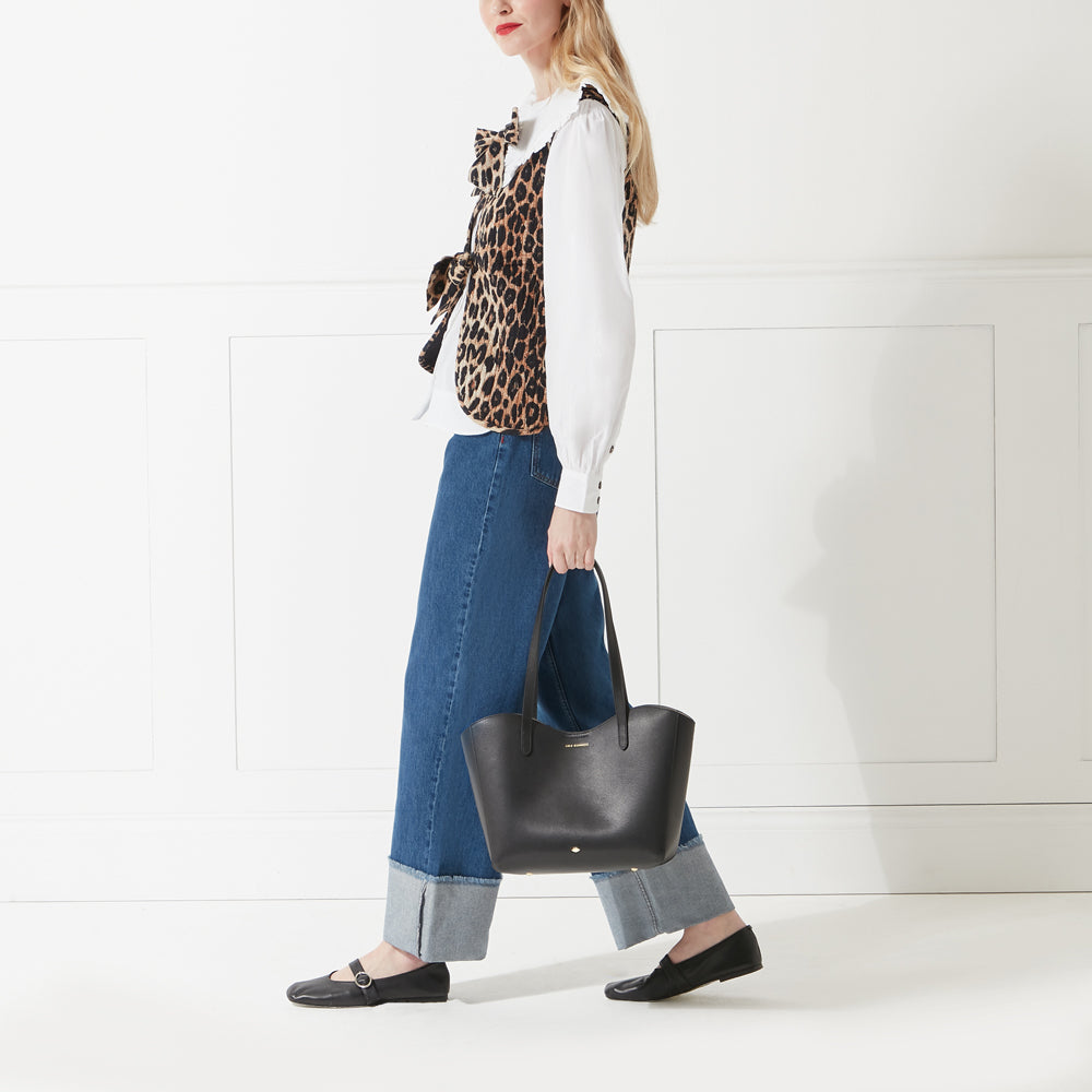 Model wearing a white shirt, jeans and a leopard print waistcoat holding a black leather tote bag with lip stud detail.