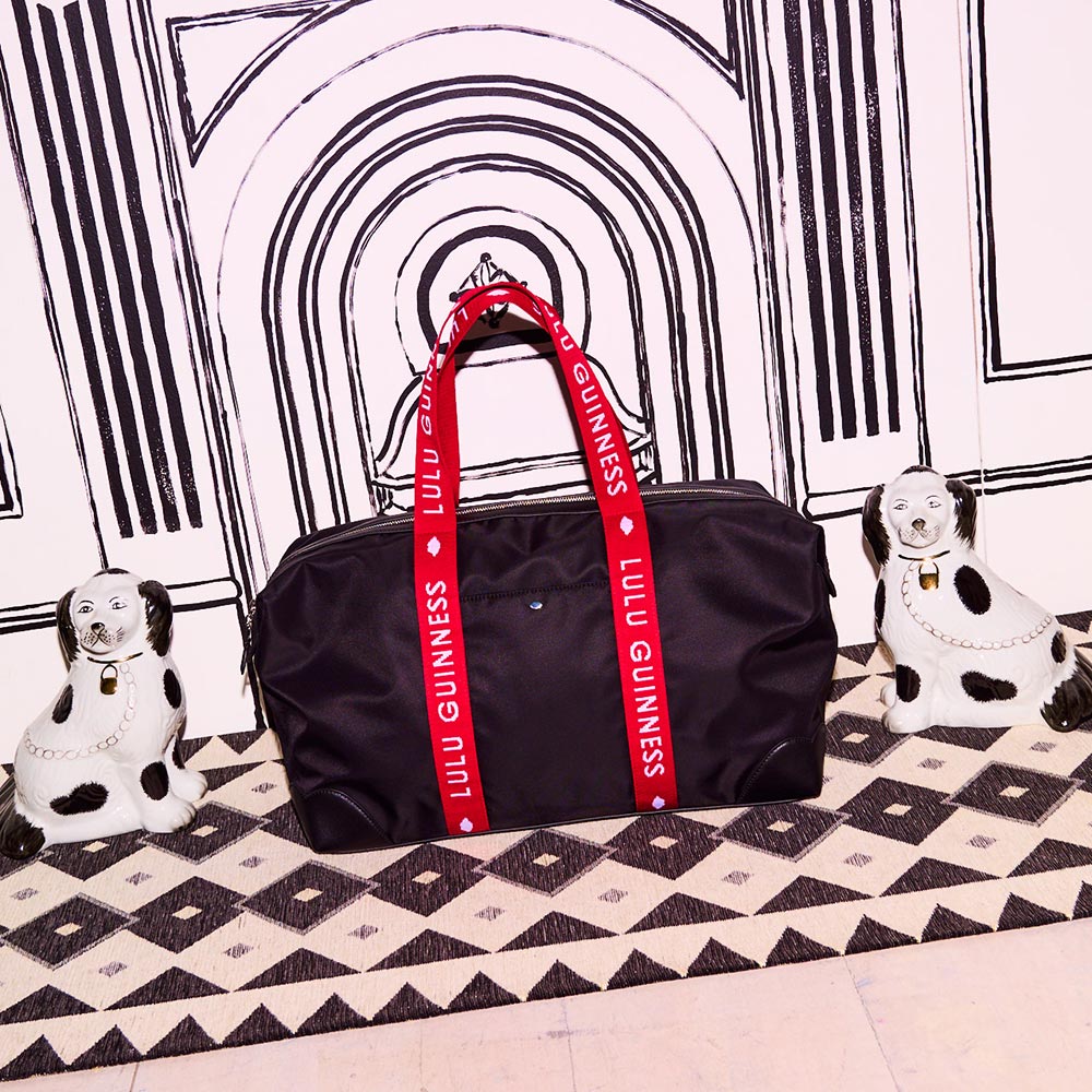 Black holdall bag with red and white branded 'Lulu Guinness' handles. Placed in front of a black and white fireplace and in-between 2 china dog ornaments. 