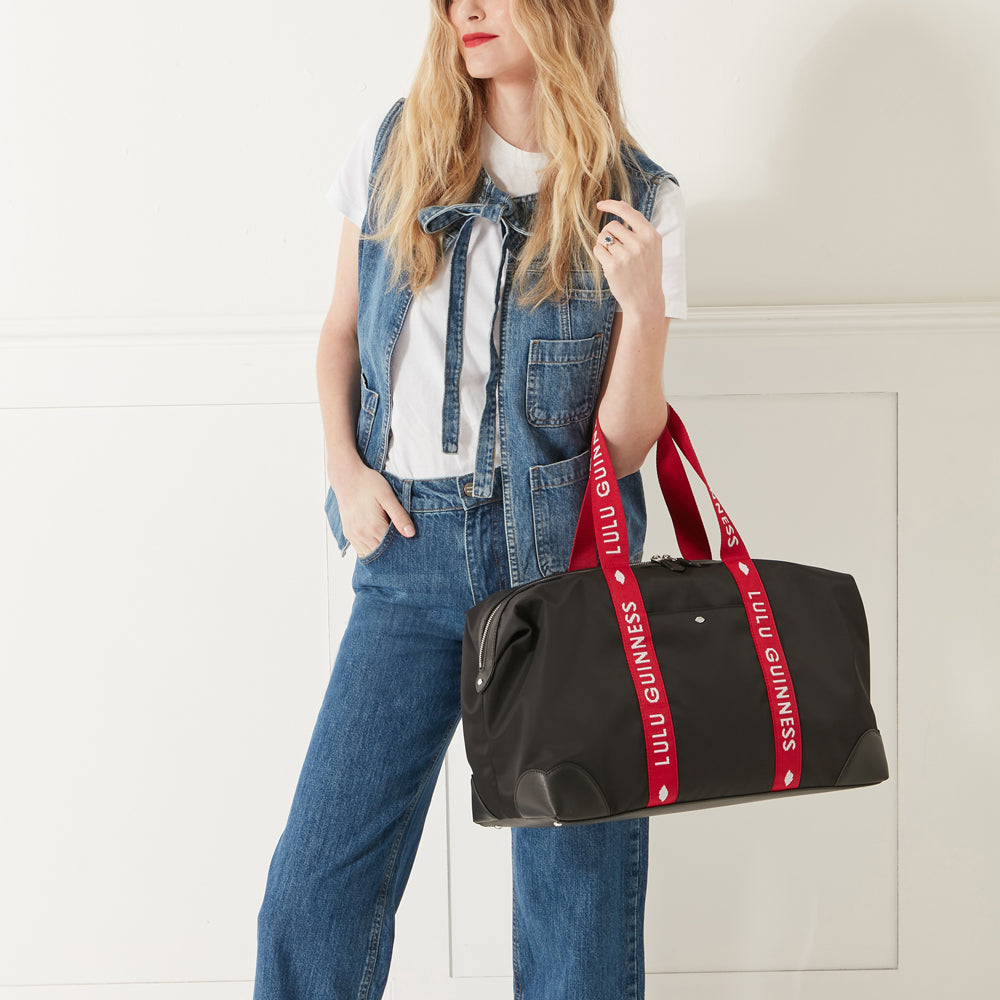 Model wearing a denim outfit, white trainers and red lipstick holding a black nylon holdall with red branded 'Lulu Guinness' handles on her arm.