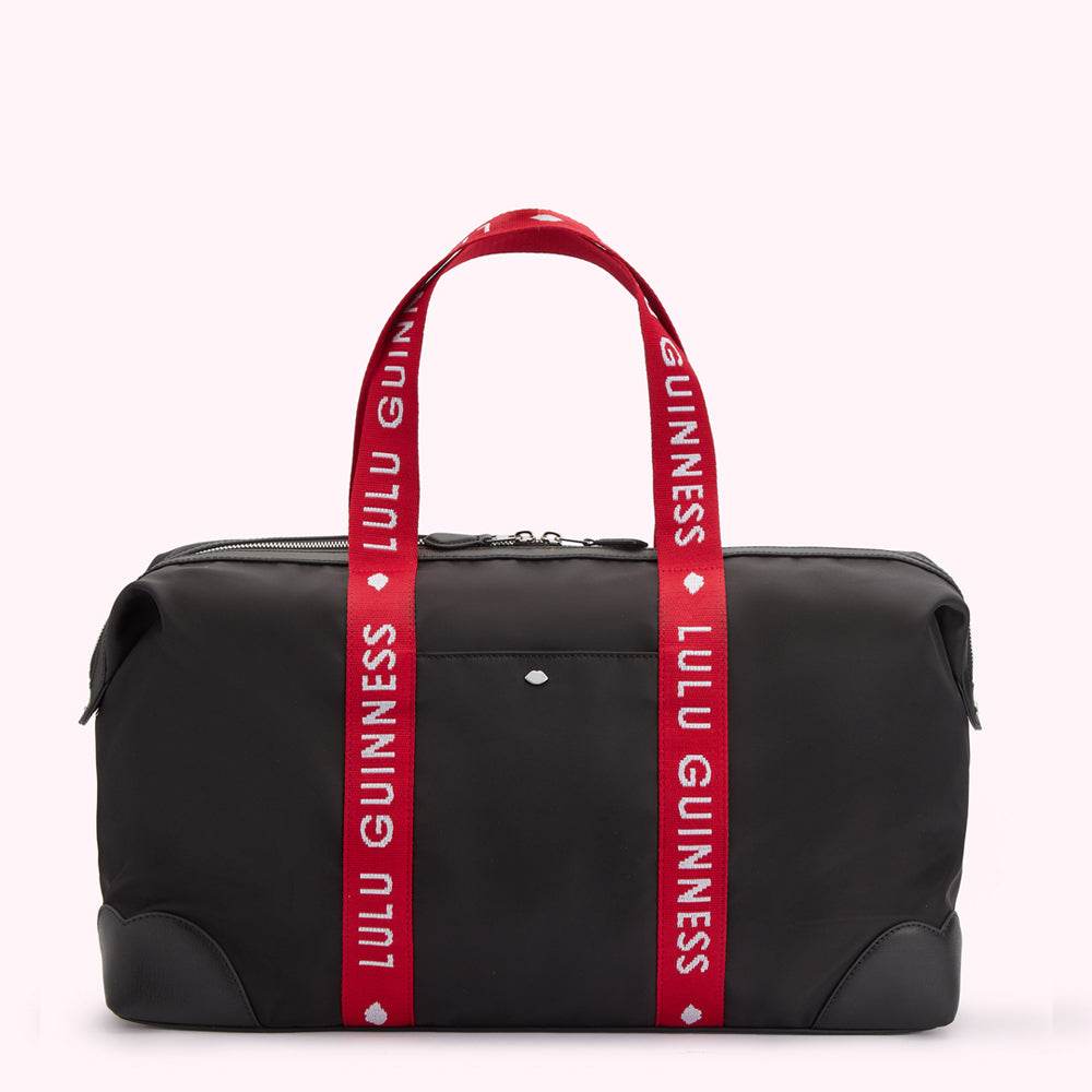 Front image of a black nylon holdall duffle bag with a red Lulu Guinness branded strap, silver metal lip pin on front pocket and curved leather corners.