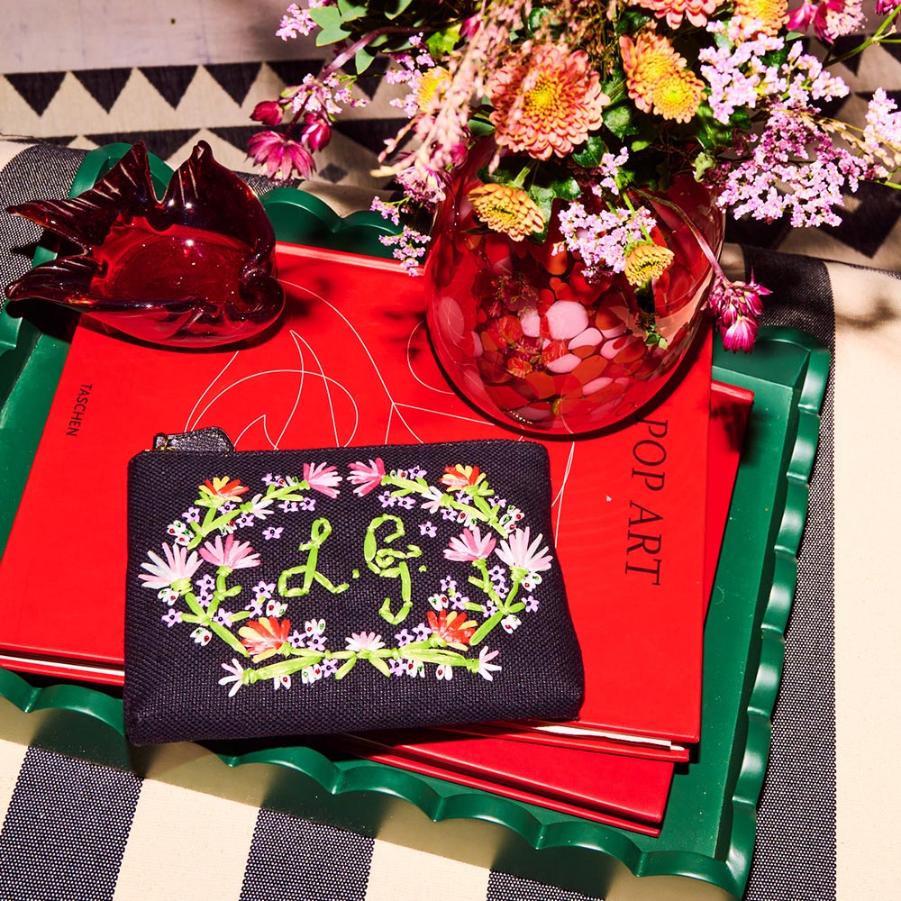 Black flower embroidered canvas pouch on top of a pile of red books next to a vase of flowers.