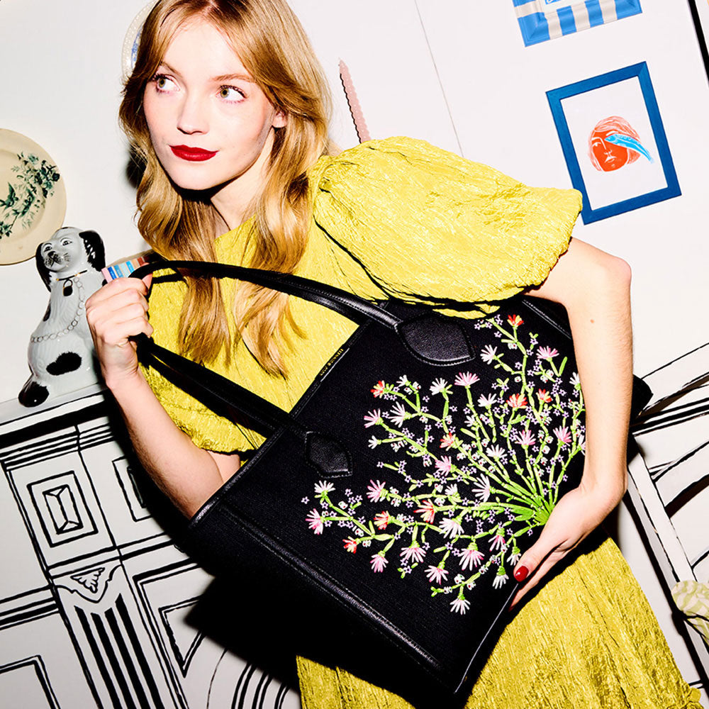 Model with blonde hair wearing a yellow dress holding a black floral embroidered tote bag.