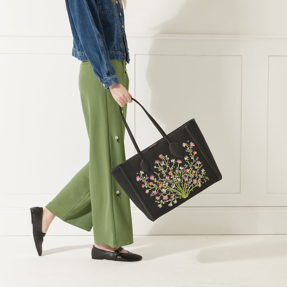 Model wearing a denim jacket, green trousers and red lipstick holding a black canvas and leather floral embroidered tote bag.