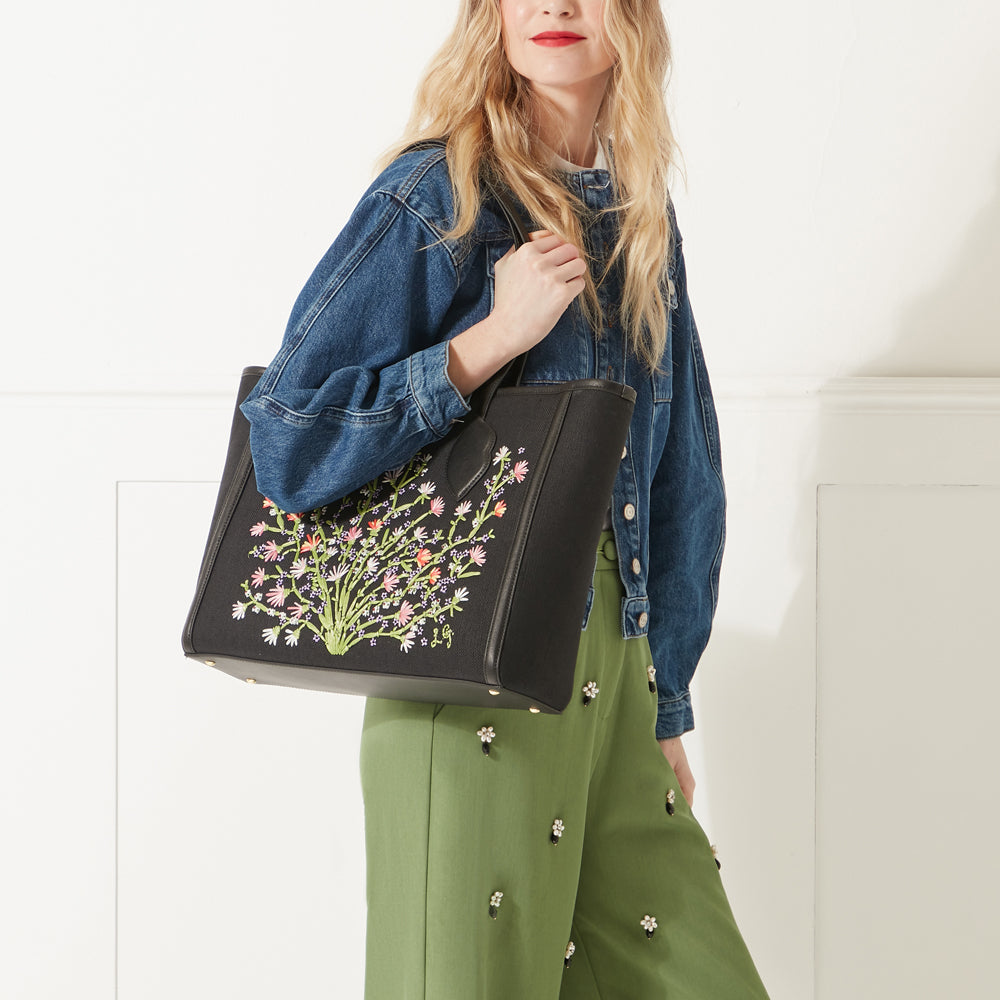 Model wearing a denim jacket, green trousers and red lipstick wearing a black canvas and leather floral embroidered tote bag on her shoulder.