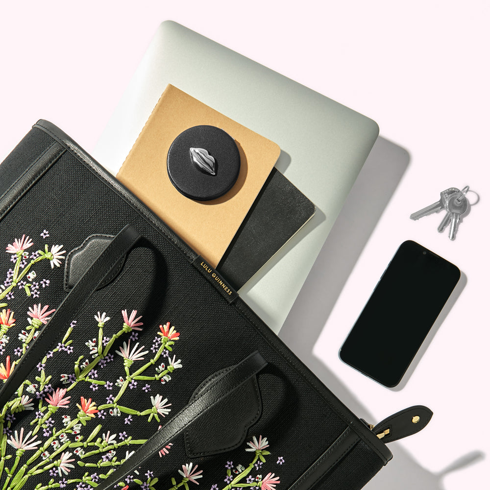 A black canvas floral embroidered tote bag handbag with a laptop, notebooks, smartphone, keys and compact mirror spilling out.