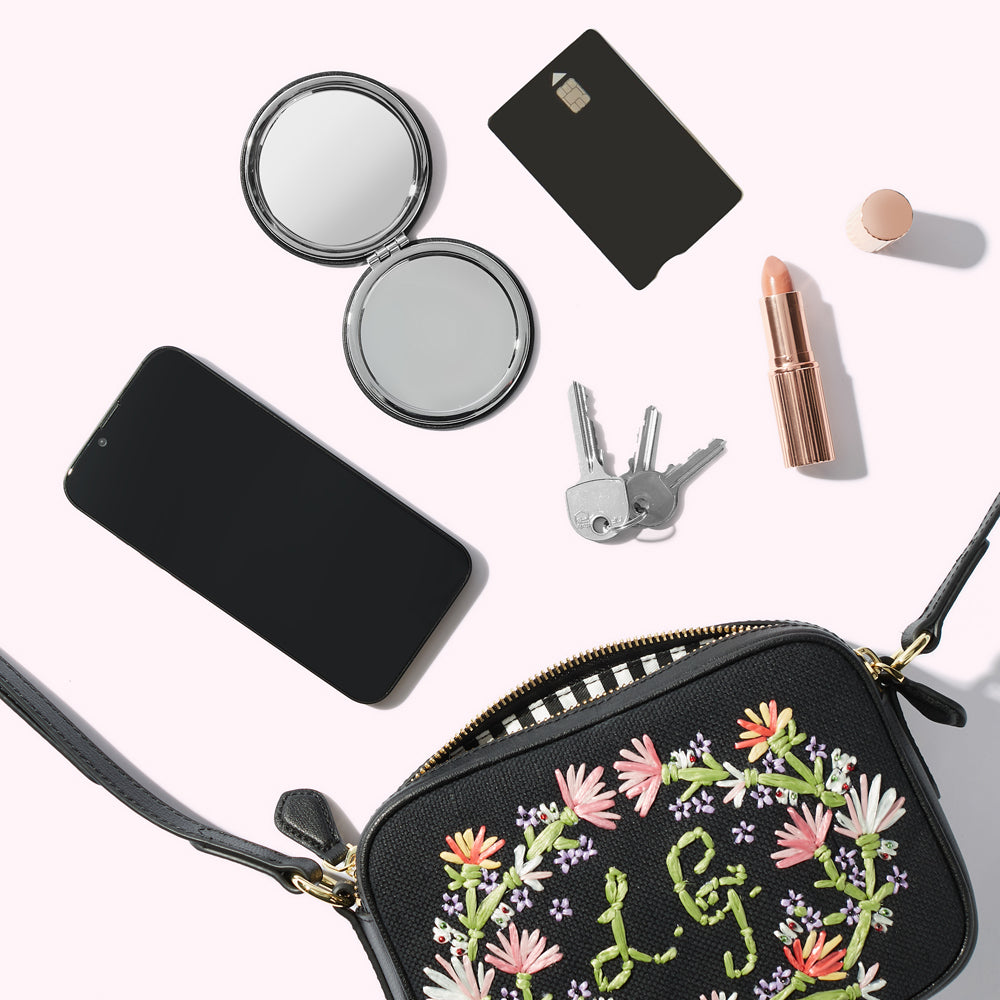 A black canvas crossbody bag with floral embroidery with a smartphone, compact mirror, keys, card and lipstick spilling out.