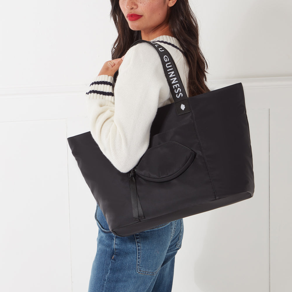 A woman in a cream sweater and jeans carries a large black tote bag with a "GUINNESS" logo on the strap. The bag has a small zippered pocket on the front.
