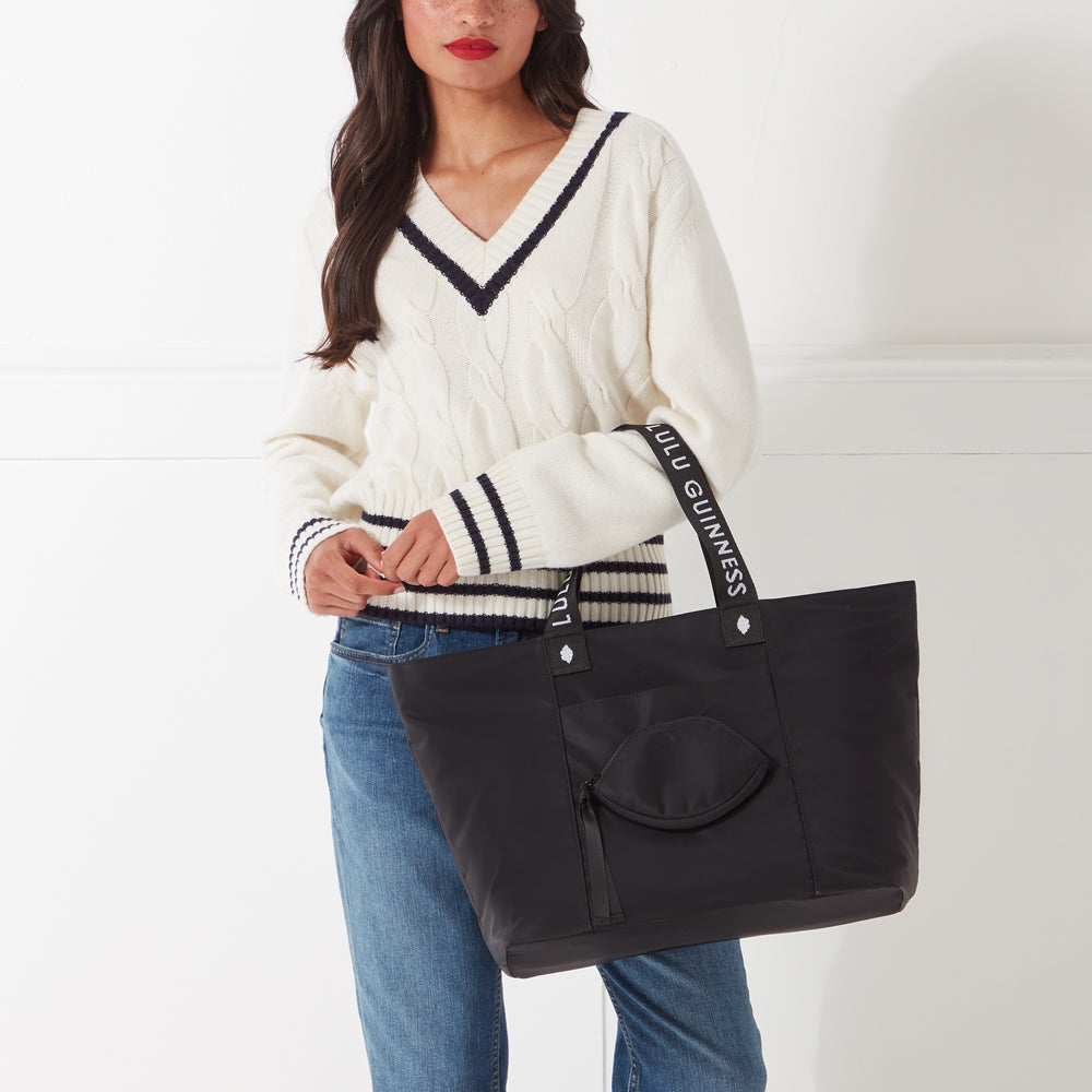 A woman in a cream sweater and jeans carries a black Lulu Guinness tote bag. The bag has a small, zipped pocket on the front and the brand name is embroidered on the straps. The background is white.
