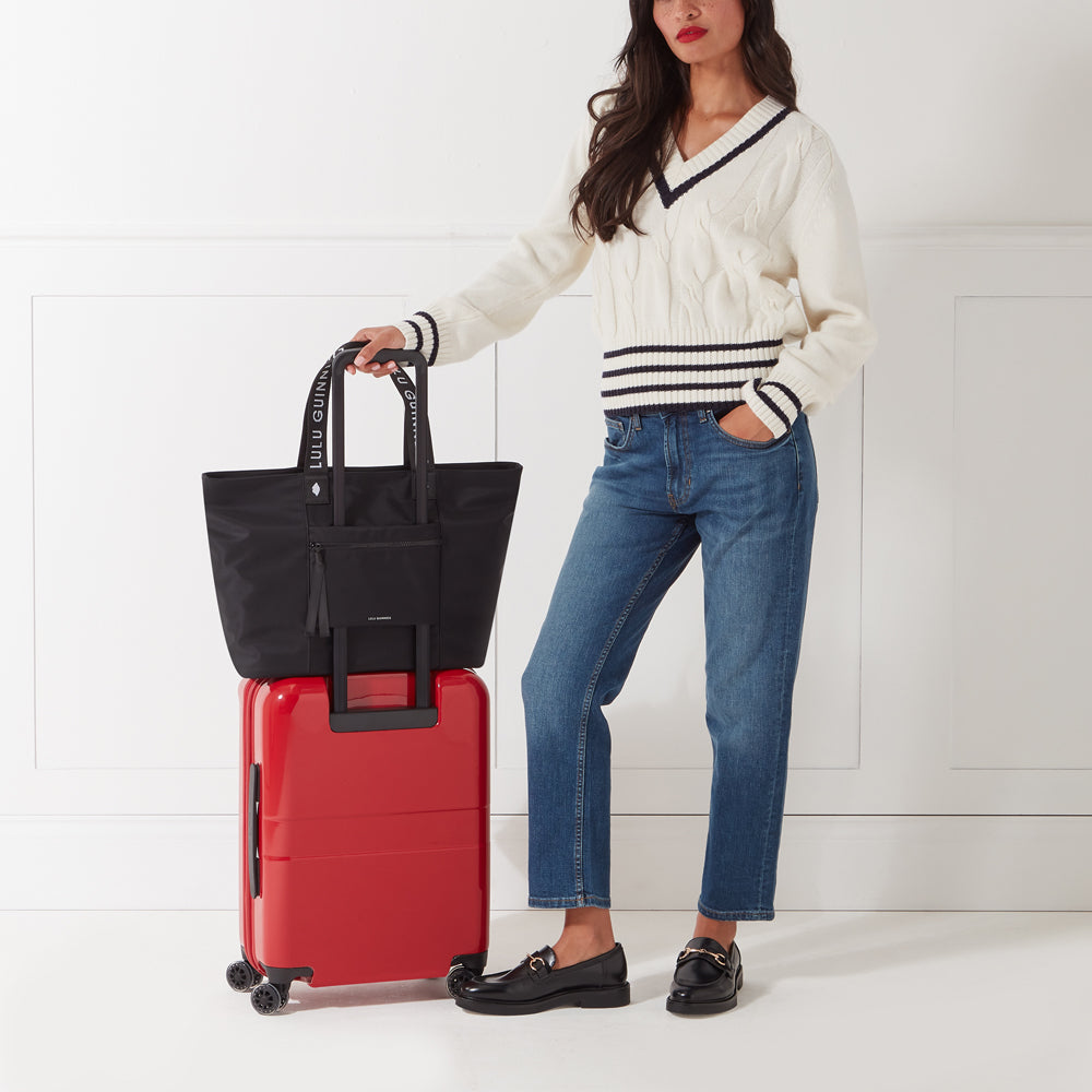 A woman in a cream sweater and jeans stands beside a red suitcase with a black tote bag on top.