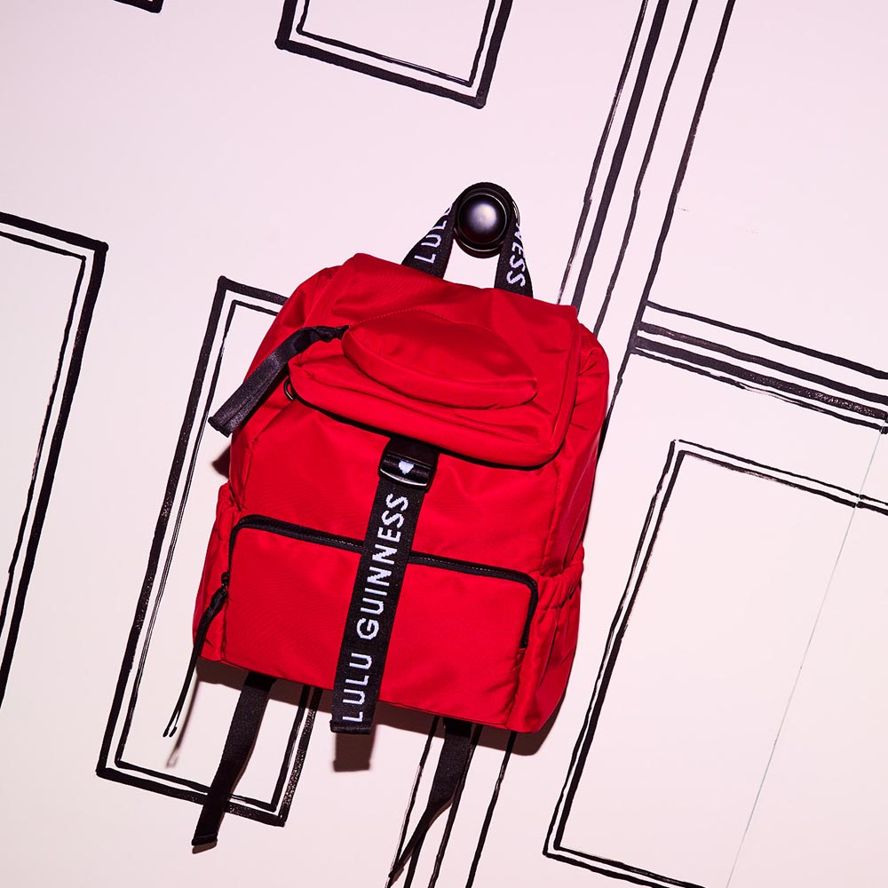 Red nylon backpack with black and with branded 'Lulu Guinness' straps hanging on a door. 