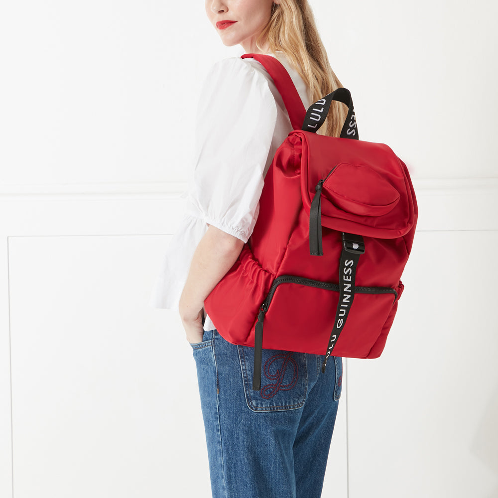 Model in a white shirt and jeans holding a red nylon backpack with 'lulu guinness' branded straps on her back.