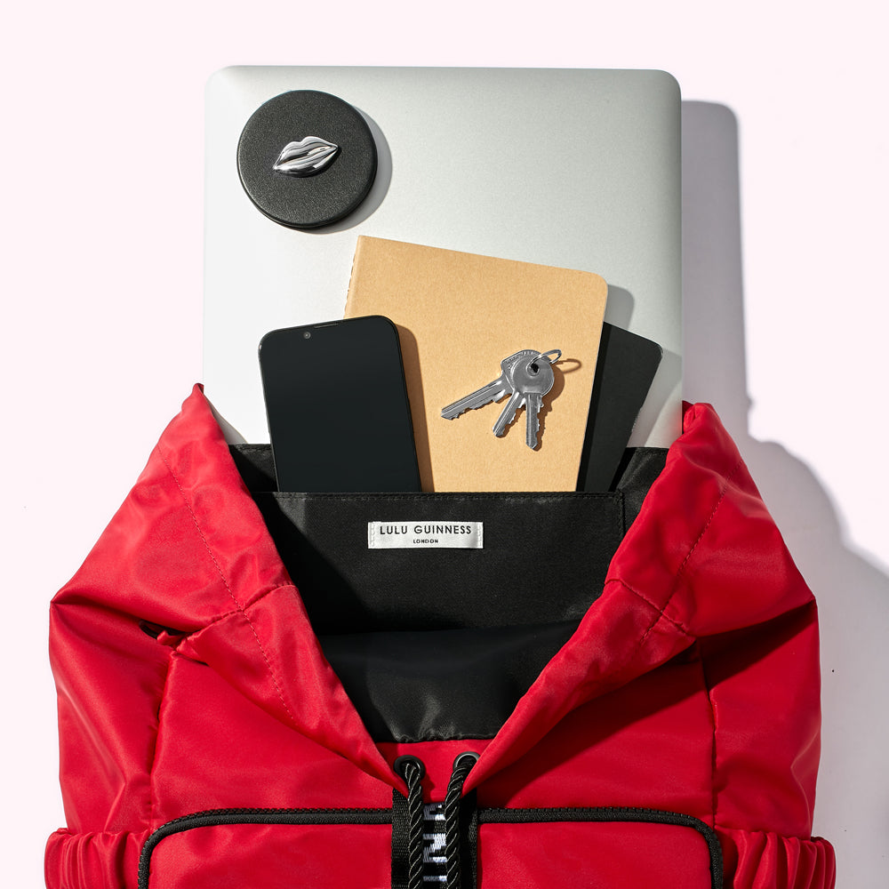 A red nylon backpack with a laptop, notebooks, smartphone, keys and compact mirror spilling out.