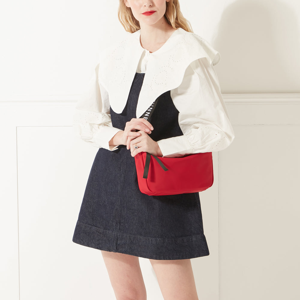 Model in a white shirt, denim dress and red lipstick wearing a red nylon shoulder bag with branded 'Lulu Guinness' shoulder strap.