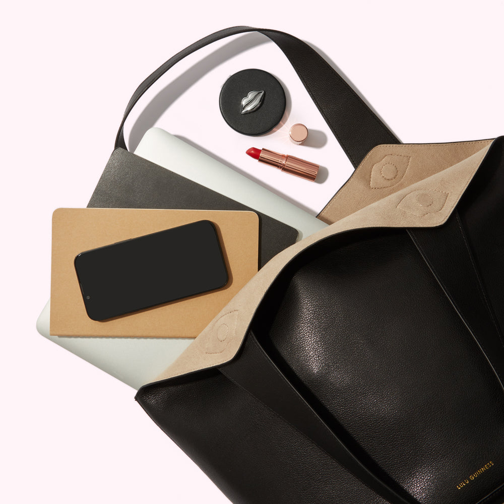 A black leather tote bag with a beige lining is open, revealing a laptop, notebooks, a smartphone, lipstick, and a compact.