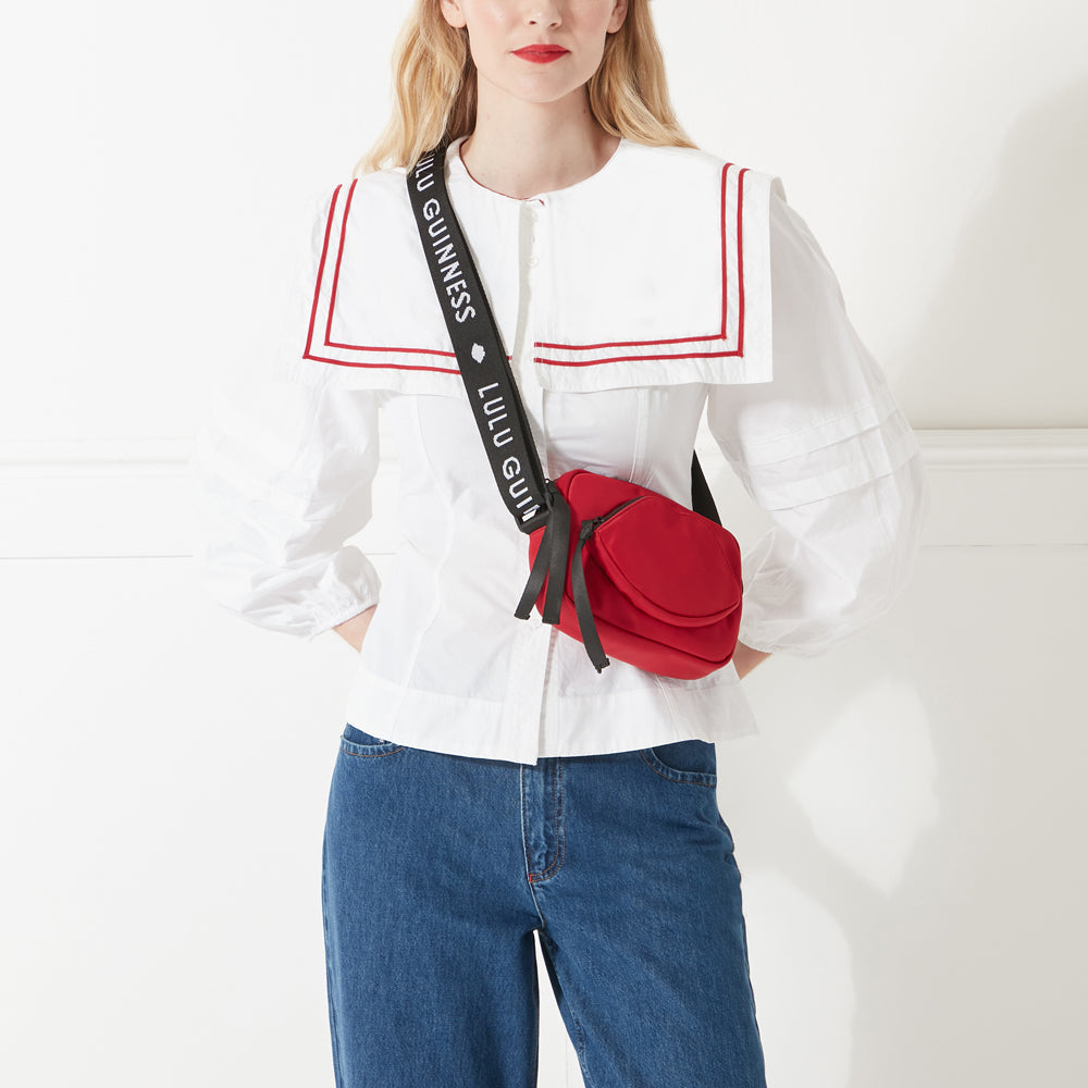 Model in white shirt and jeans wearing a red nylon crossbody bag with a lip shaped pocket and black branded 'Lulu Guinness' strap. 