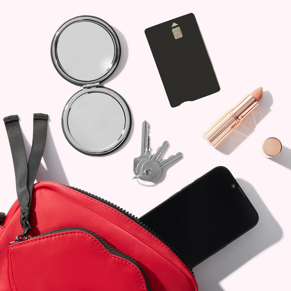 A red nylon crossbody bag with a smartphone, keys, compact mirror, card and lipstick spilling out.