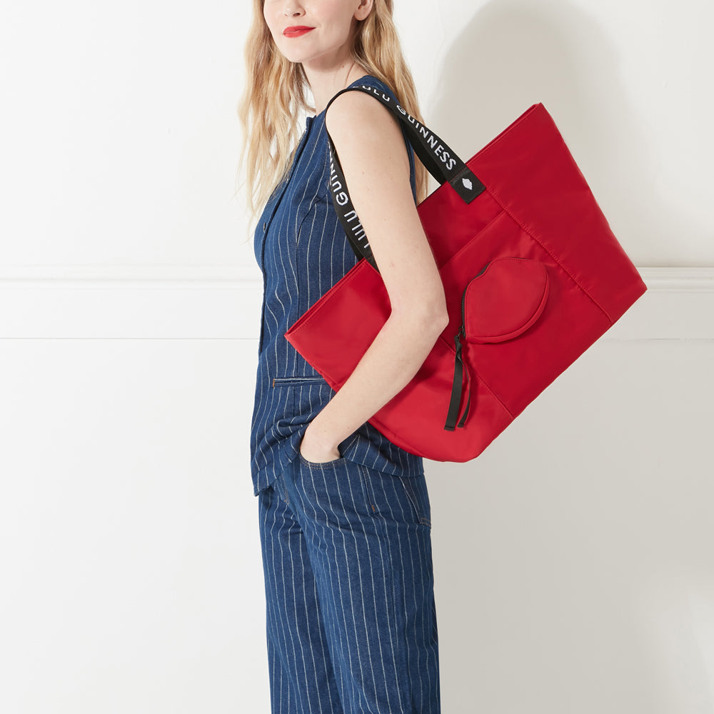 Model in denim outfit wearing a red nylon tote bag with lip shaped pocket and branded 'Lulu Guinness' straps on her shoulder.