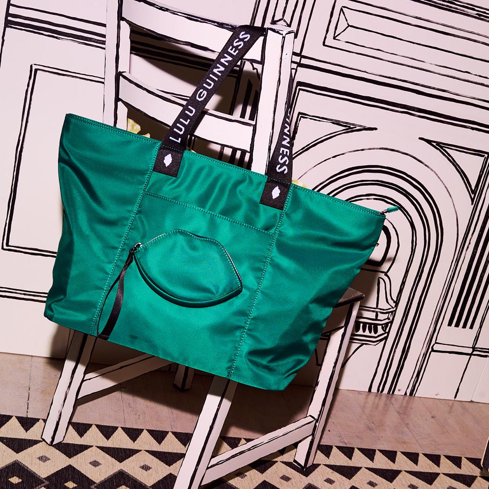 Emerald green nylon tote bag with a lip shaped pocket on the front and black 'Lulu Guinness' branded straps hanging from a black and white chair.