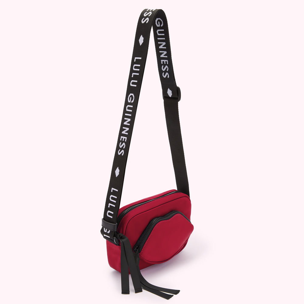 Red crossbody bag with a lip-shaped detail and black strap featuring repeating "Lulu Guinness" branding.
