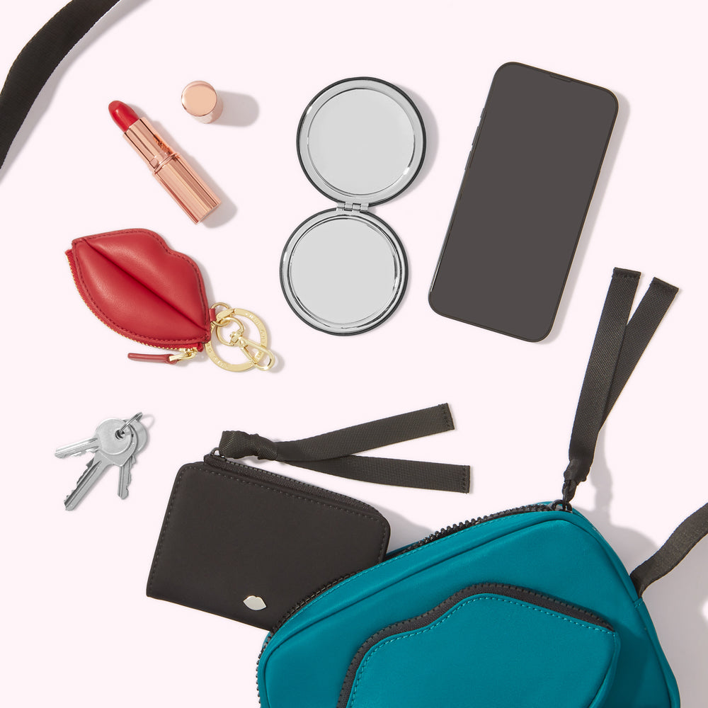 A teal crossbody bag spills its contents: a black cardholder, keys, a red lip-shaped keychain, a compact mirror, lipstick, and a smartphone. All items are arranged on a white background.
