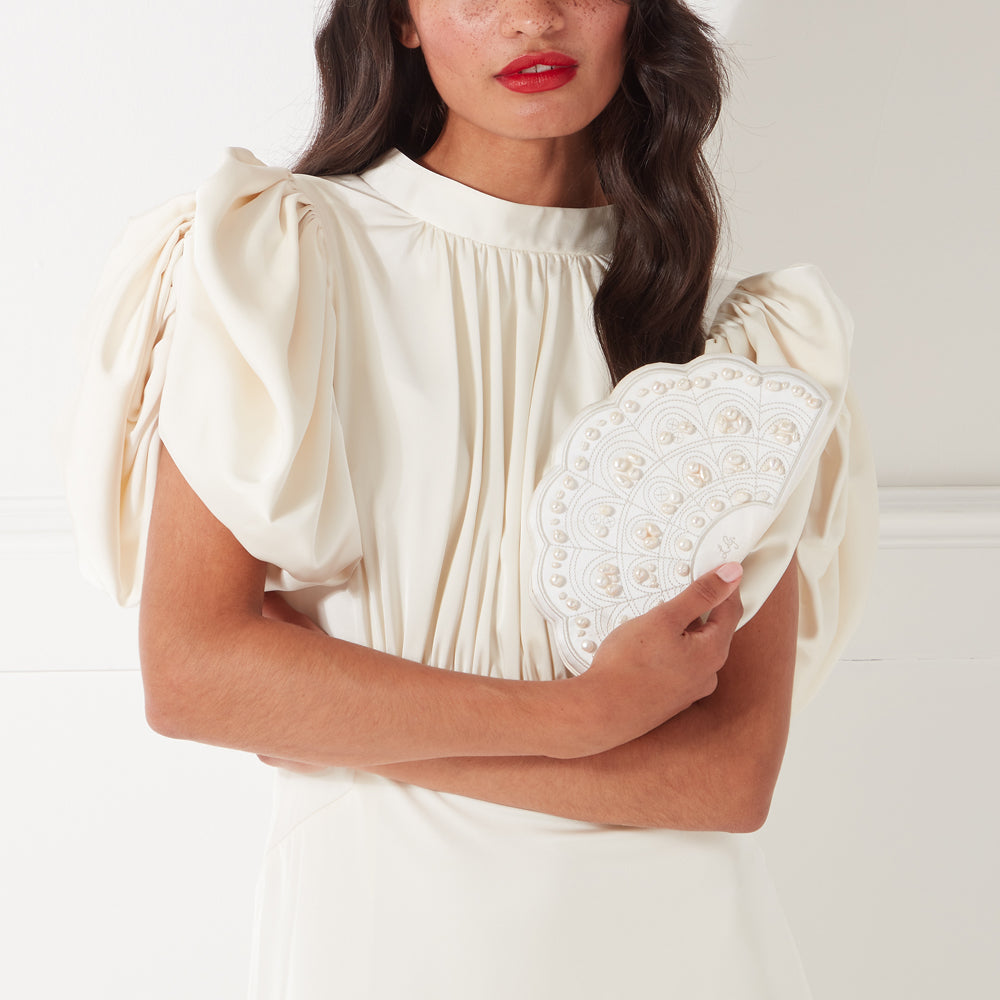 Woman in off-white dress holding a pearl-embellished fan-shaped clutch.
