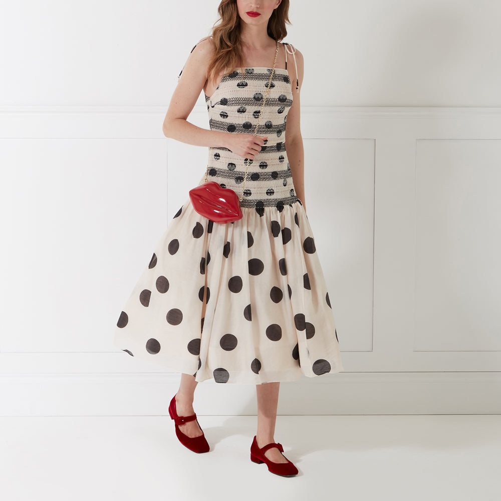 Woman in cream polka dot dress and red shoes carrying a red lip-shaped purse against a white background.
