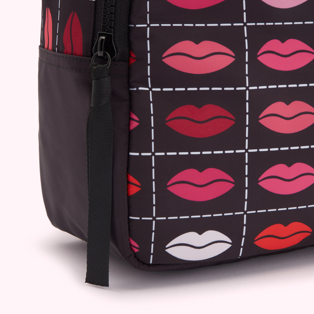 Close-up of a black backpack with a repeating pattern of lips in various shades of red and pink on a dark background. The backpack has a black zipper and a black pull-tab.
