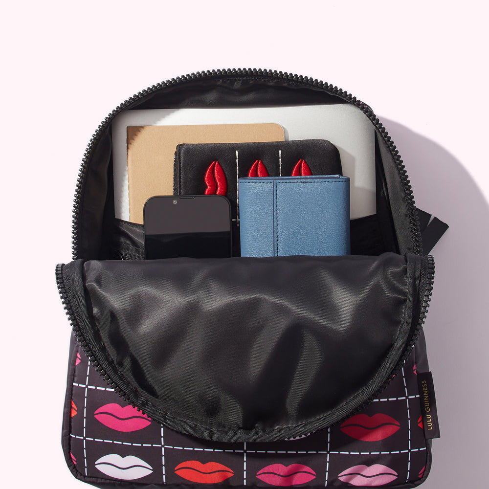 Open Lulu Guinness backpack with a black and red lip print, containing a laptop, notebooks, phone, and wallet.
