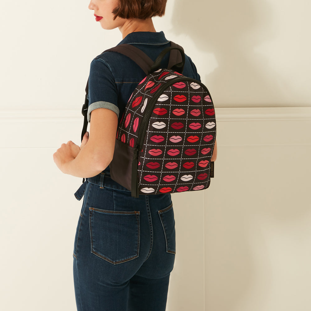 A woman in dark denim overalls models a black backpack with a repeating pattern of red and pink lips.