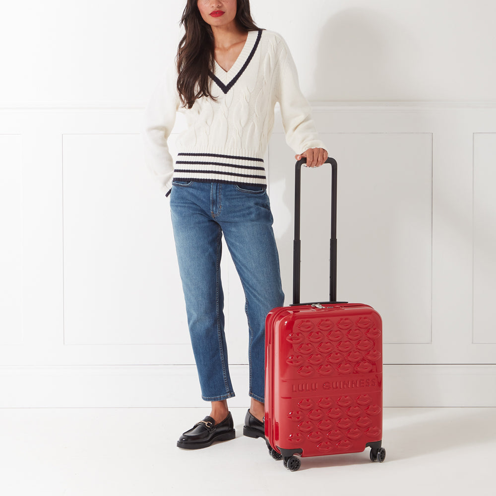 Woman in cream sweater and jeans standing next to a red Lulu Guinness carry-on suitcase featuring a lip design.
