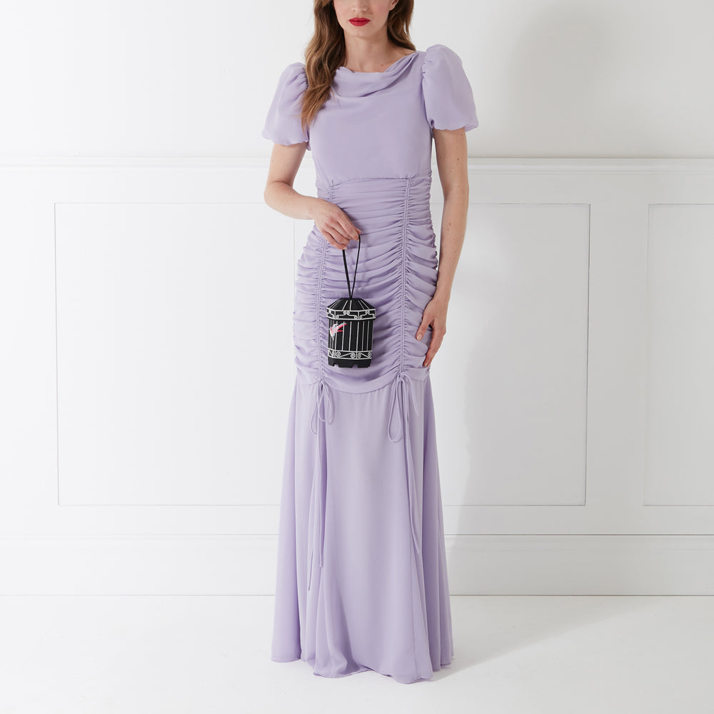 Woman in lavender dress holding a black birdcage-shaped purse against a white background.
