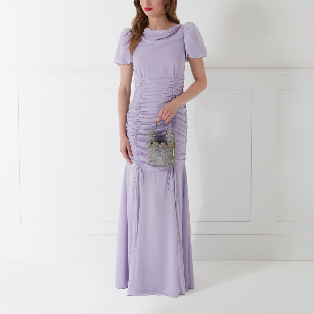 A woman in a lavender dress holds a silver castle-shaped handbag.
