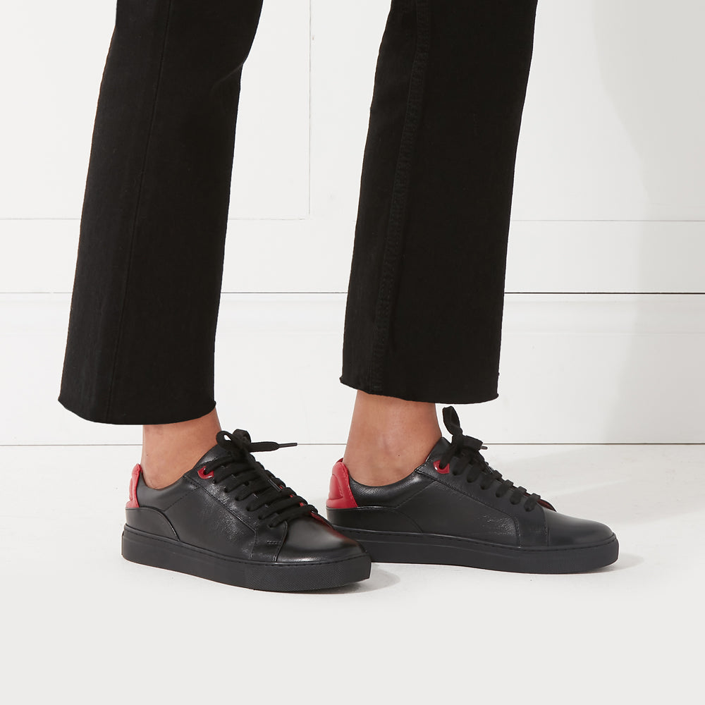 Black leather sneakers with red accents, worn by a model in black jeans against a white background.
