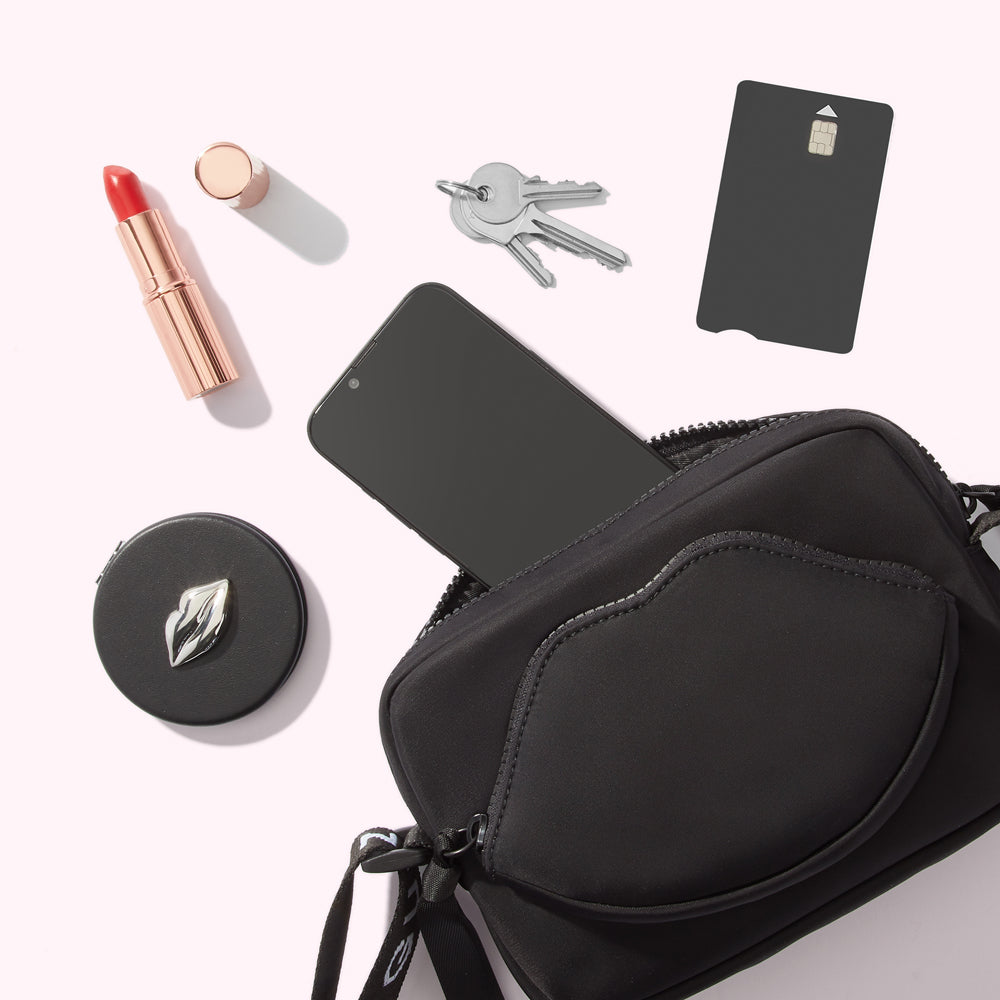 A black crossbody bag with a lip-shaped pocket lies open, displaying its contents against a white background: a smartphone, lipstick, compact mirror, keys, and credit card.