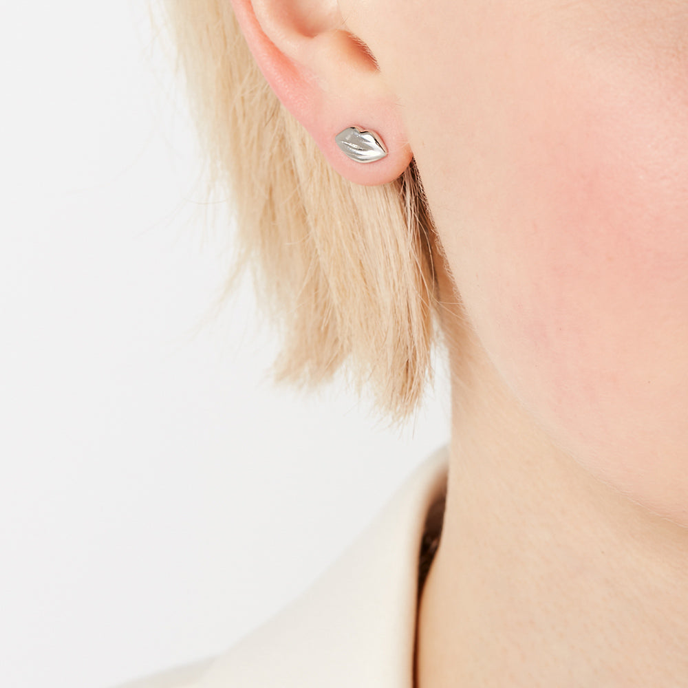 Close-up of a person with blonde hair wearing silver lip-shaped stud earrings against a white background.
