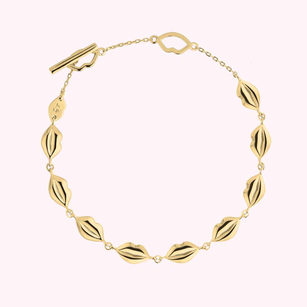 Gold chain bracelet featuring linked lip-shaped charms and a toggle clasp, displayed against a light pink background.