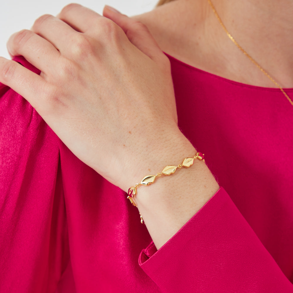 Close-up of a woman's wrist wearing a gold bracelet with leaf-shaped charms against a vibrant fuchsia background.
