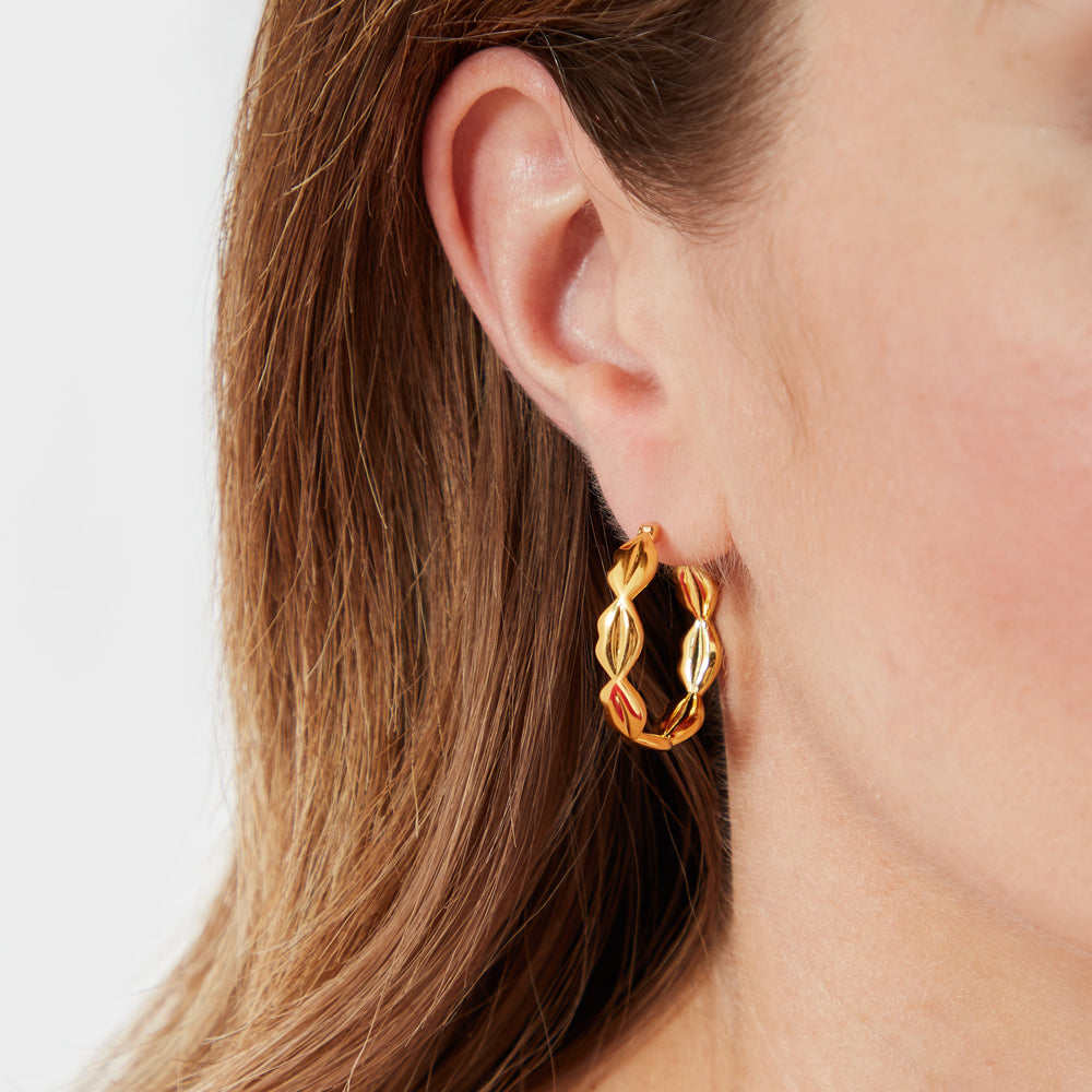 Close-up of a woman's ear wearing gold hoop earrings with a textured, leaf-like design against a white background.
