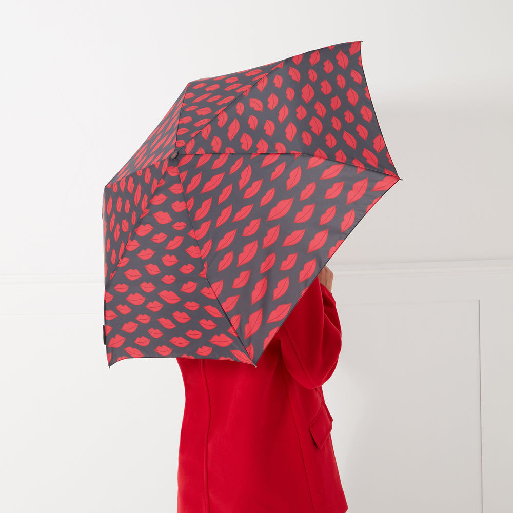 A woman in a red coat holds a dark grey umbrella with a pattern of red lips printed on it.
