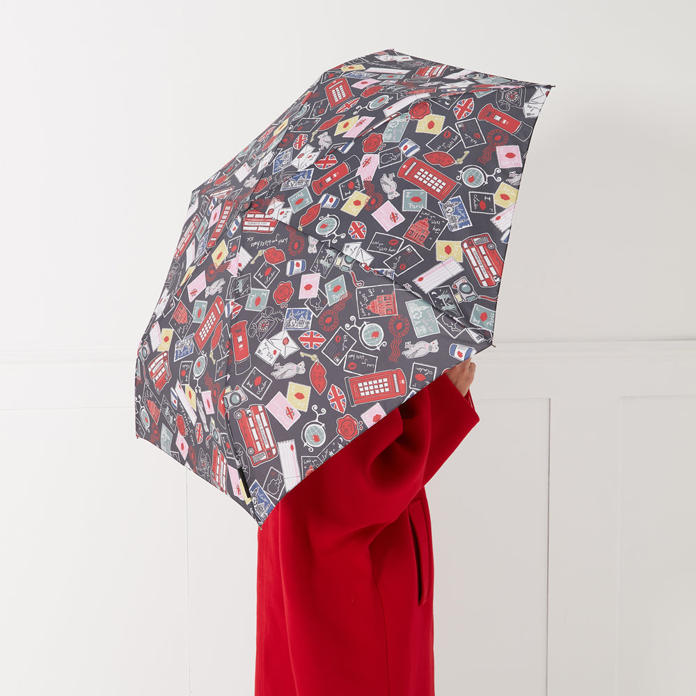 Compact umbrella with a dark background and a colourful pattern of postage stamps, red lips, London landmarks and other related images. A person wearing a red coat holds the umbrella.
