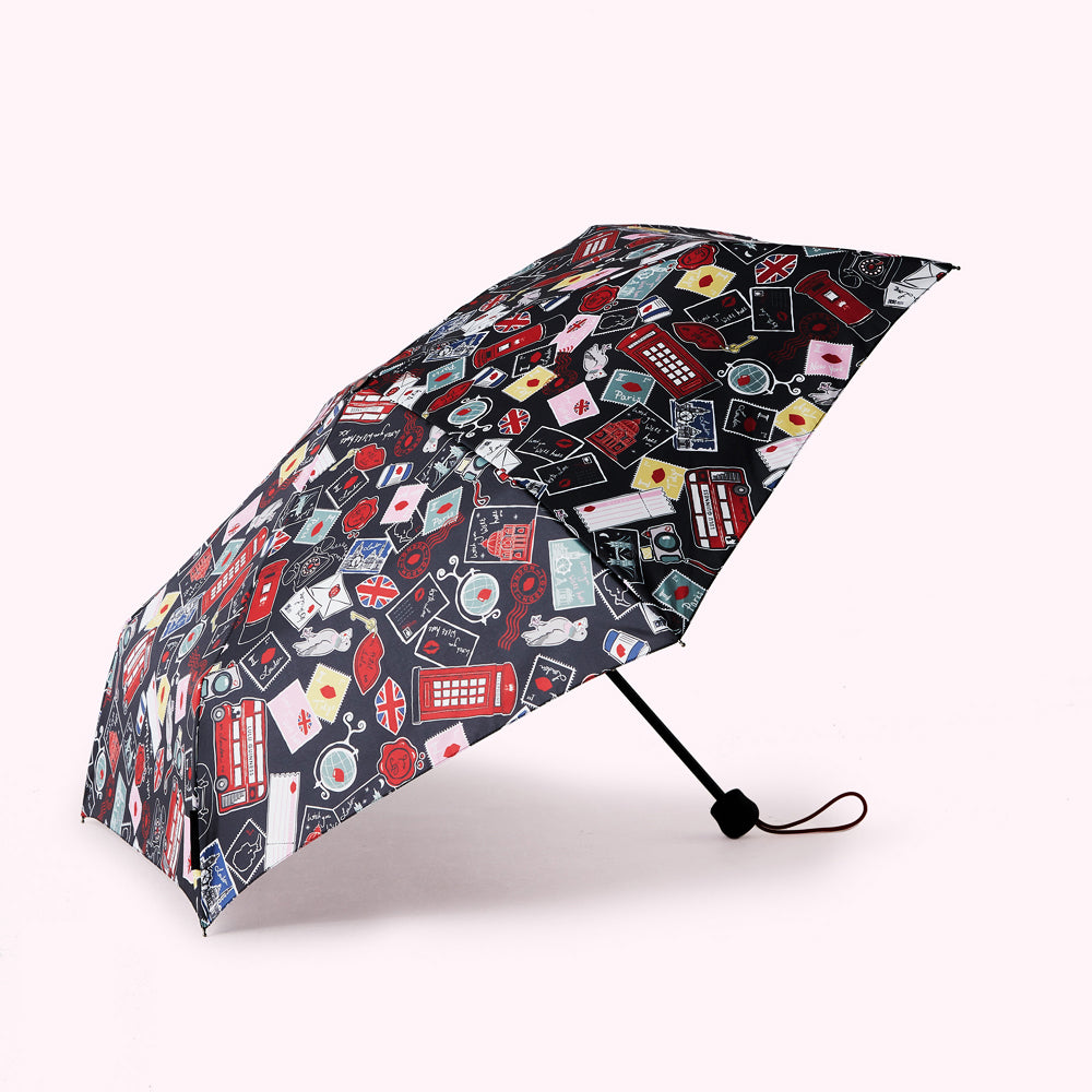 Compact travel umbrella with a dark background and a colourful pattern featuring London landmarks, postmarks, and other travel-themed imagery.
