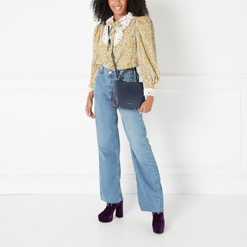 Woman in floral blouse, jeans, and purple platform shoes, carrying a navy blue handbag against a white background.
