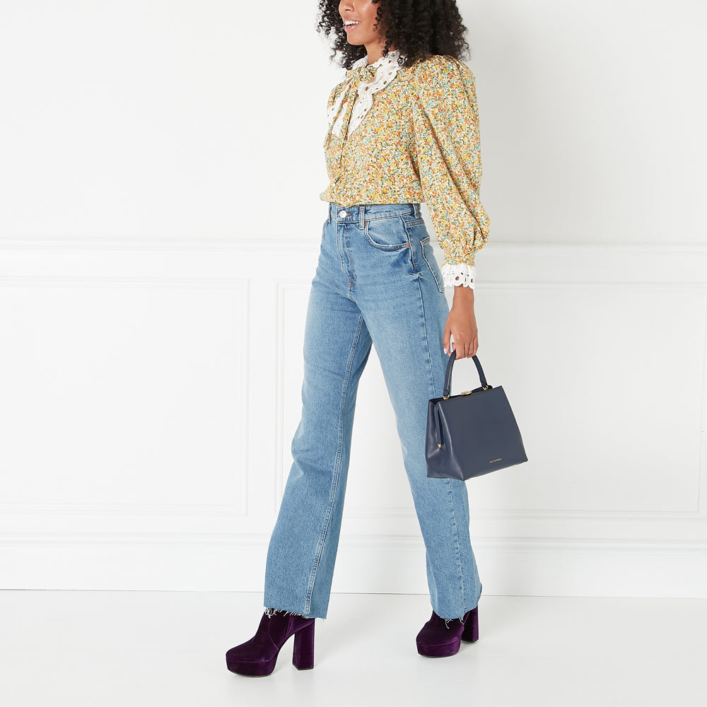 A woman with curly hair wears a floral blouse, light wash jeans, and purple platform ankle boots. She carries a dark blue top-handle handbag. The background is white.
