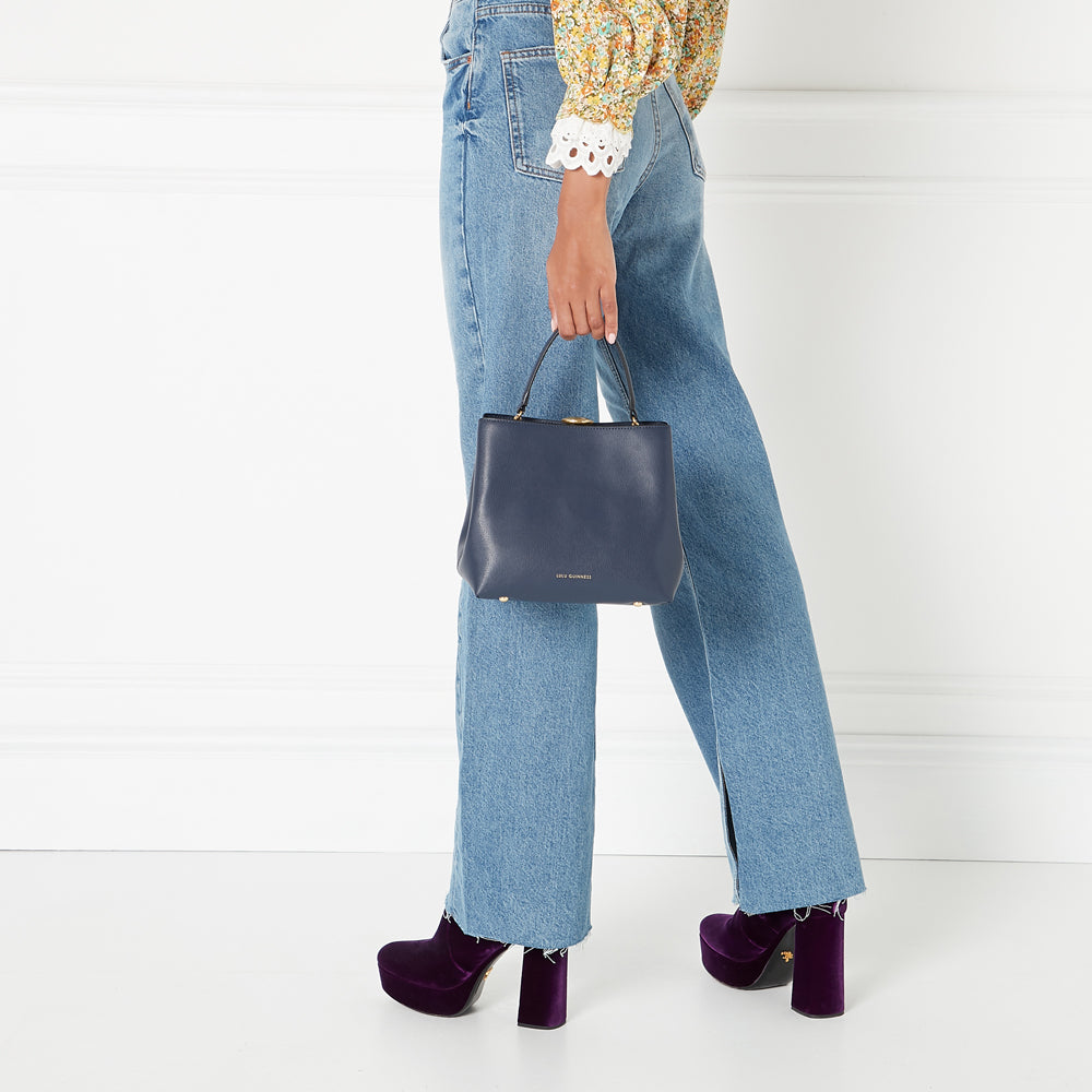 A woman in light-wash jeans and purple platform shoes holds a navy blue Lulu Guinness bag against a white background.