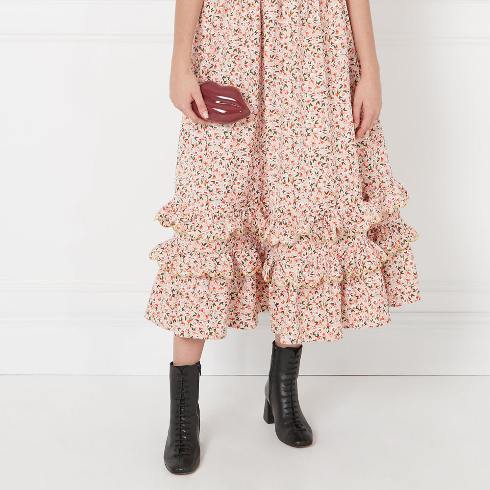 A woman in a floral dress and black ankle boots holds a dark red lip-shaped clutch.